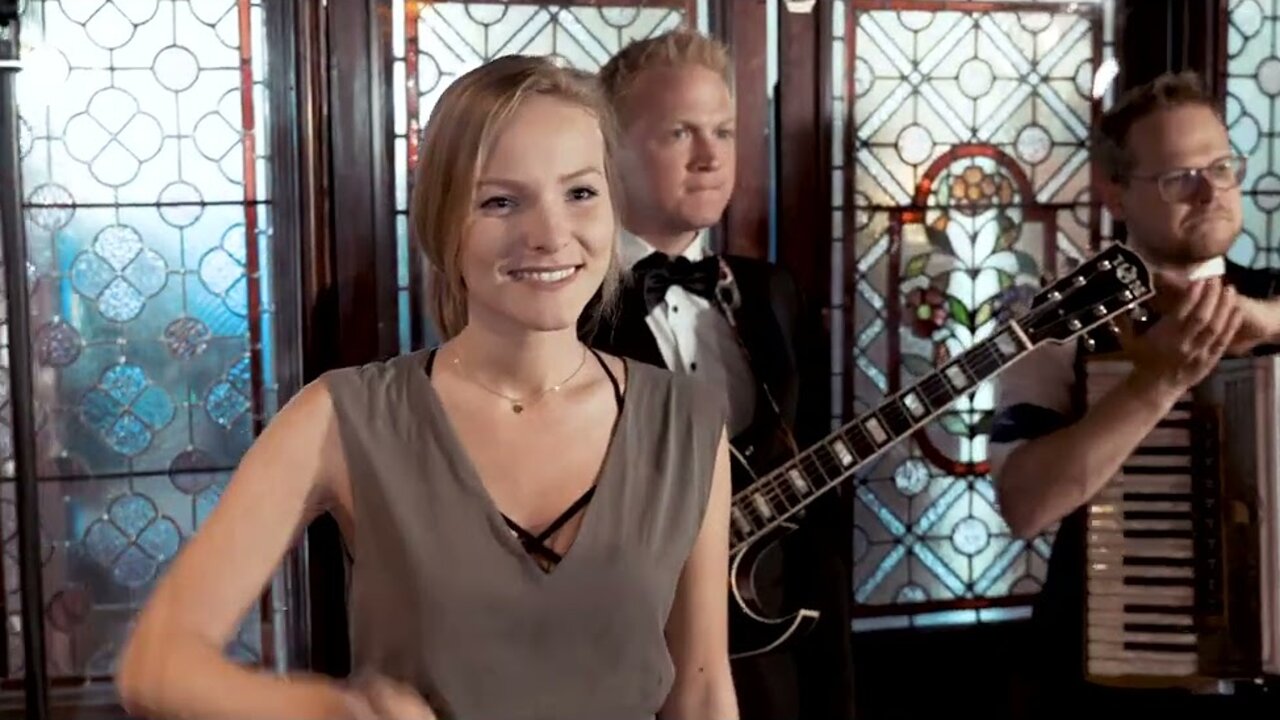 Standbild aus dem Musikvideo „Humpa Humpa“: eine Sängerin der Fäaschtbänkler, dahinter Gitarrist und Akkordeonspieler vor bunten Bleiglasfenstern