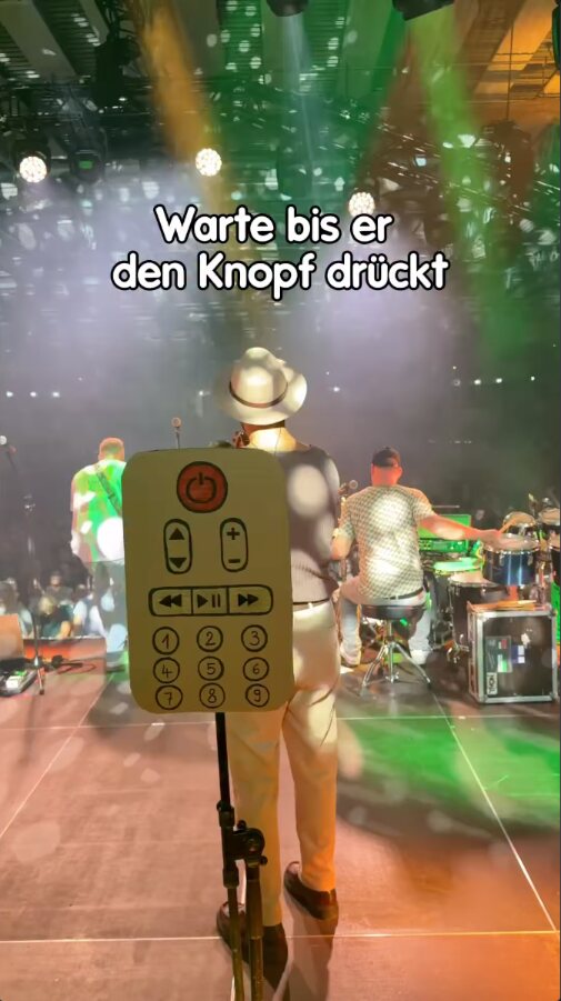 Reel der Fäaschtbänkler: ein Musiker mit einem Pappschild in Form einer Fernbedienung steht auf der Bühne vor Band und Publikum, darüber der Text „Warte bis er den Knopf drückt“