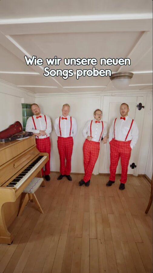 Reel der Fäaschtbänkler: vier Musiker in roten Hosen und Hosenträgern stehen in einer holzvertäfelten Stube neben einem Klavier, darüber der Text „Wie wir unsere neuen Songs proben“