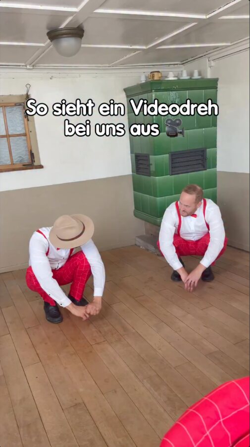 Reel der Fäaschtbänkler: zwei Musiker hocken beim Videodreh auf dem Holzboden einer Stube mit grünem Kachelofen, darüber der Text „So sieht ein Videodreh bei uns aus“